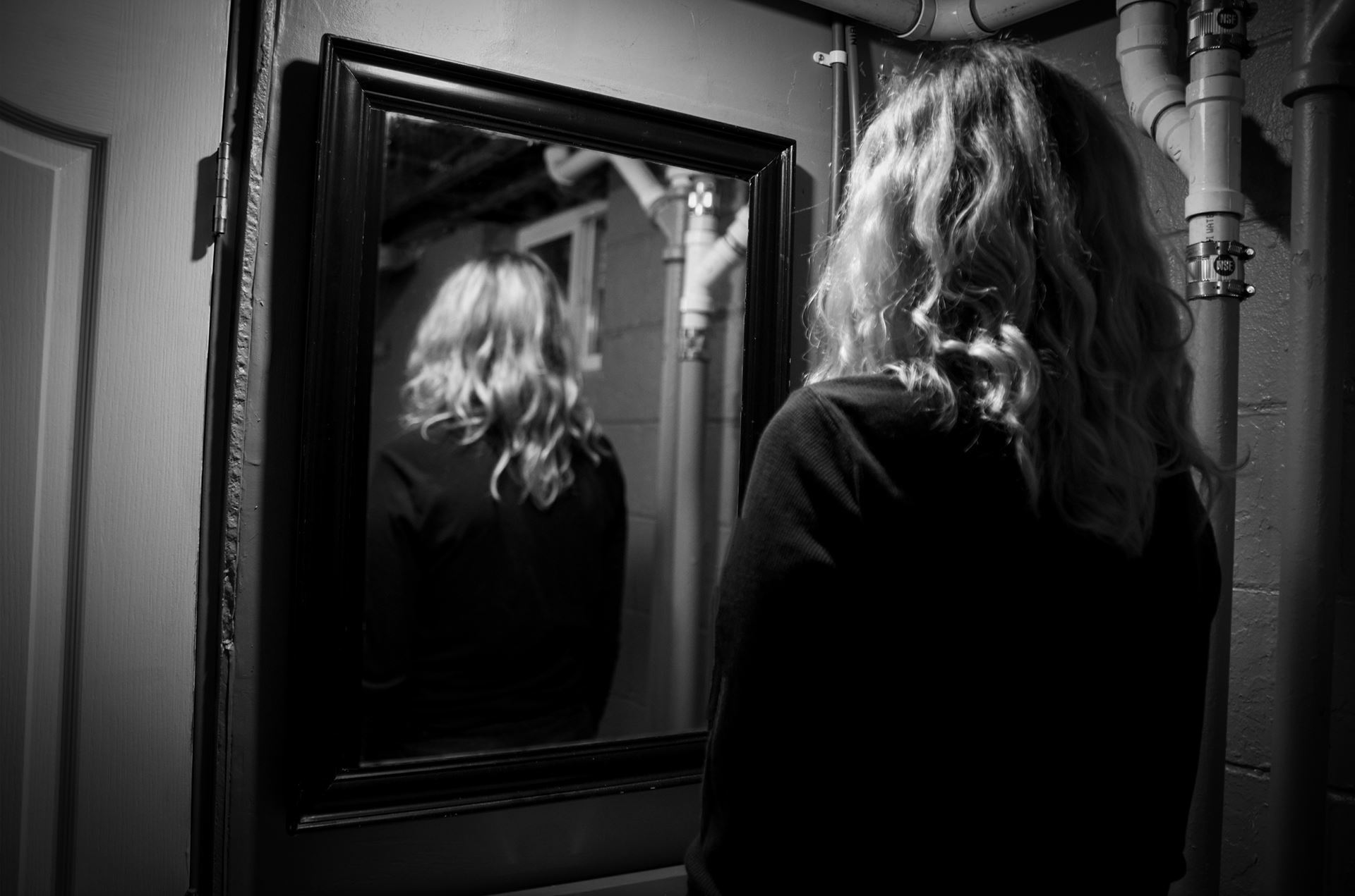 Black and white photo, person facing away at a mirror, which also shows them facing away. Annie Suckow, “Reflections I,” digital photo, 8.5"x11"